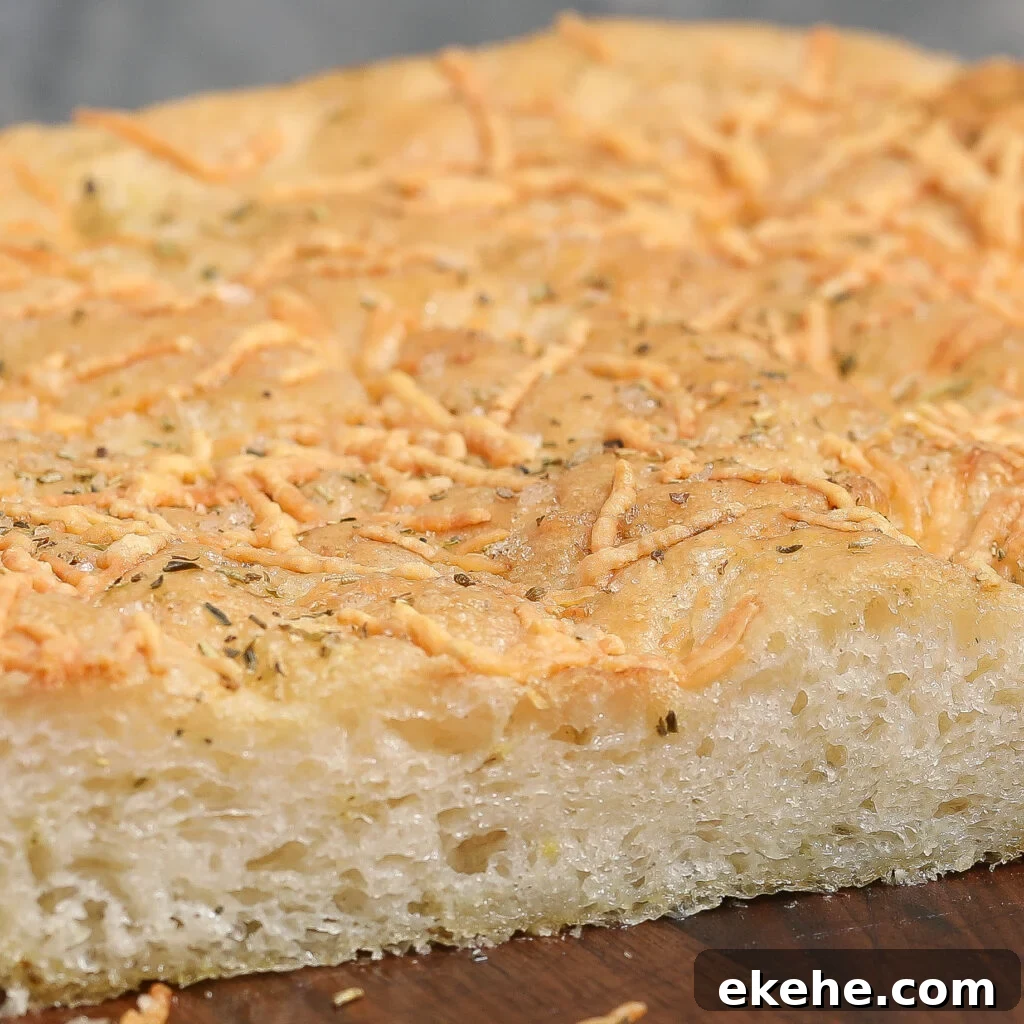 A close-up of a thick slice of Parmesan Herb Focaccia, topped with shredded cheese and herbs, showing a golden, slightly crisp crust and a soft, airy interior.