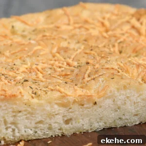 A close-up of a thick slice of Parmesan Herb Focaccia, topped with shredded cheese and herbs, showing a golden, slightly crisp crust and a soft, airy interior.