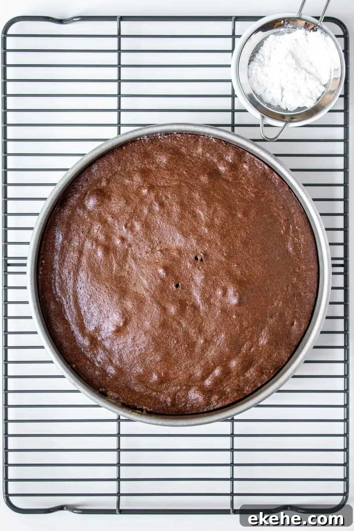 Cake in pan cooling on rack with powdered sugar in small seive next to it.