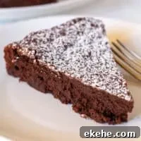 Piece of bittersweet chocolate cake with powdered sugar on white plate with fork.