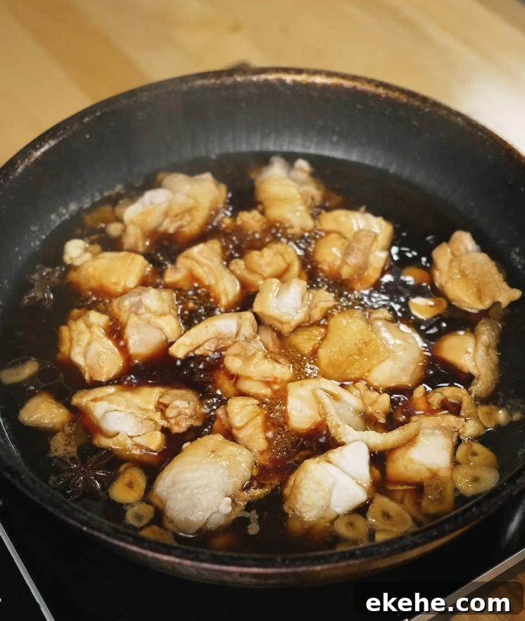Chunks of chicken thigh are shown simmering in a dark soy-based sauce with visible slices of ginger and garlic, absorbing deep umami flavors.