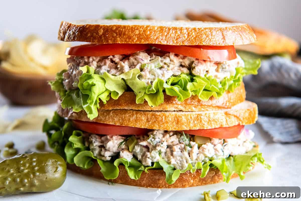 Dill pickle tuna salad sandwich layered with lettuce and tomato on sliced bread, served with potato chips and a pickle on the side.