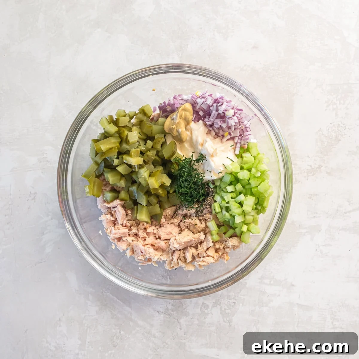 Ingredients for dill pickle tuna salad added to a glass bowl, including albacore tuna, chopped dill pickles, celery, red onion, mayonnaise, Dijon mustard, fresh dill, and black pepper before mixing.