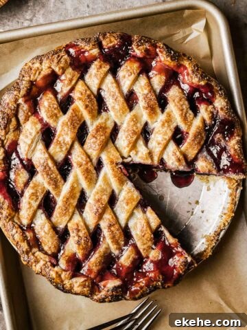 A golden brown lattice topped baked cherry rhubarb pie on a parchment lined baking sheet.