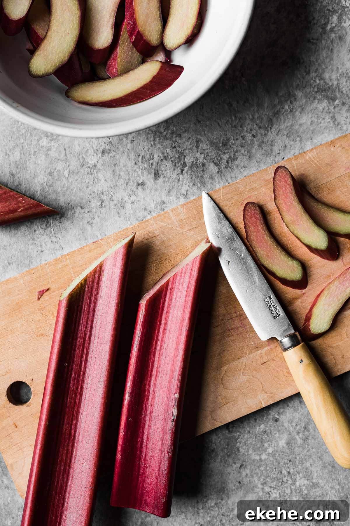 A wooden cutting board with reddish pink rhubarb stalks cut on the bias. A wooden handled paring knife rests nearby.