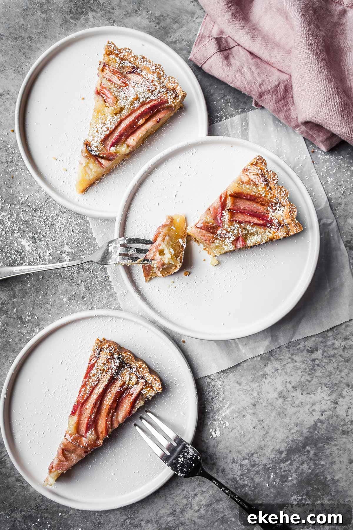 Three slices of fruit tart on white ceramic plates with two forks resting on the plates and a pink napkin nearby.