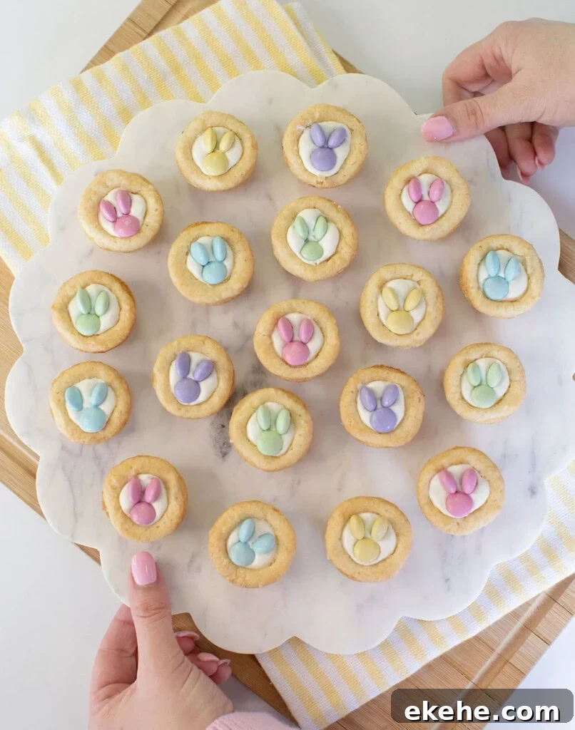 A person holds a round marble tray with 19 cookies topped with pastel-colored candy decorations shaped like bunny paw prints. The tray is on a striped yellow and white cloth over a wooden board.