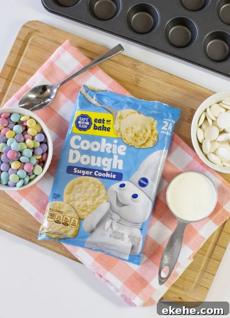 A bag of Pillsbury sugar cookie dough is on a wooden board with a red-and-white checkered towel, surrounded by pastel chocolate candies, white chocolate wafers, and a bowl of milk near a muffin pan.