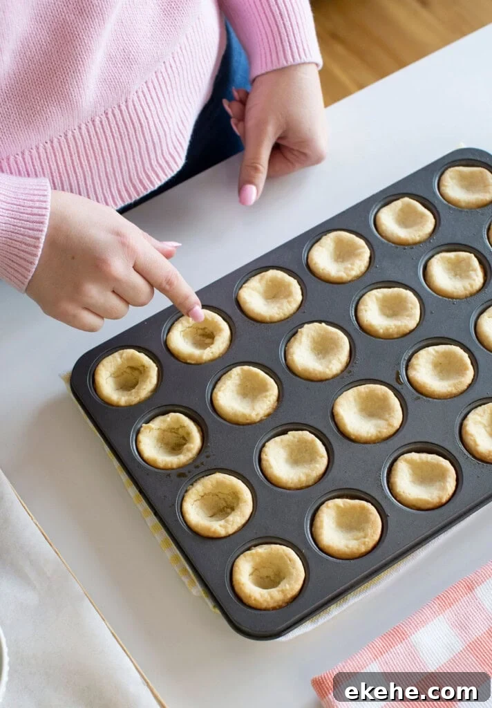 A person in a pink sweater touches a mini tart shell in a muffin tin filled with empty, baked tart shells on a white countertop.