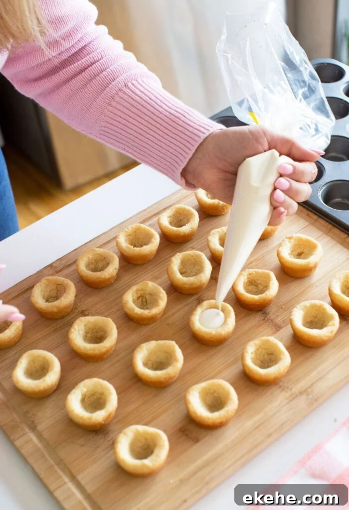 A person uses a piping bag to fill hollowed-out cupcake bites arranged on a wooden board; a muffin tin is visible nearby.