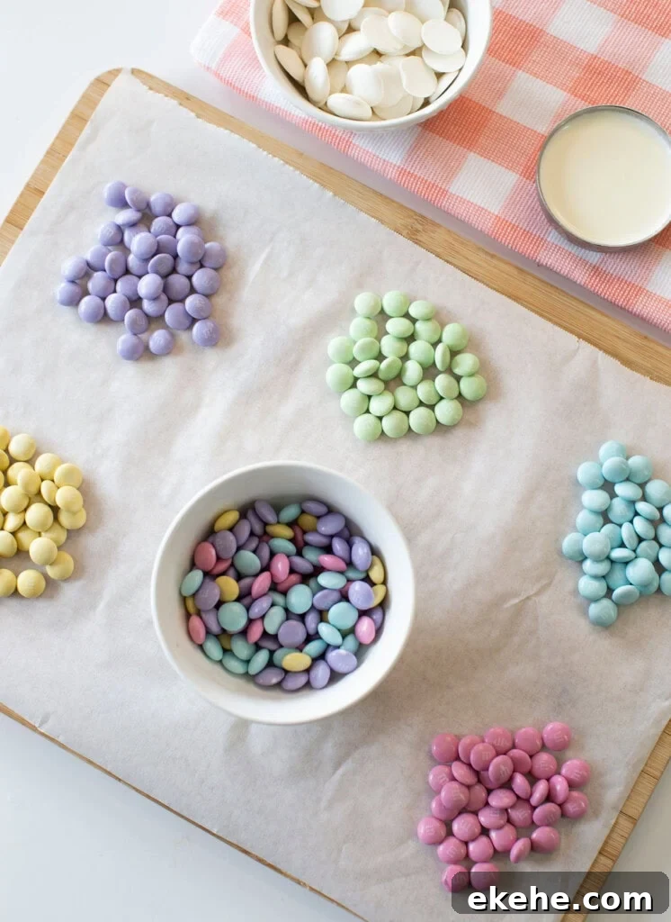 Six small piles of colorful candy-coated chocolates are arranged on parchment paper, with a bowl of mixed candies in the center. Nearby are a bowl of white chocolate wafers and a cup of liquid on a pink-checked cloth.