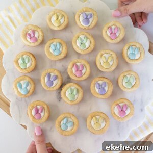 A person holds a round marble tray with 19 cookies topped with pastel-colored candy decorations shaped like bunny paw prints. The tray is on a striped yellow and white cloth over a wooden board.