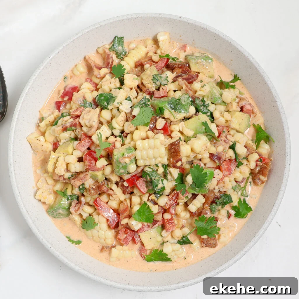 A bowl of creamy corn and avocado salad with bacon, chopped red bell pepper, cilantro, and a light orange dressing sits on a white surface.