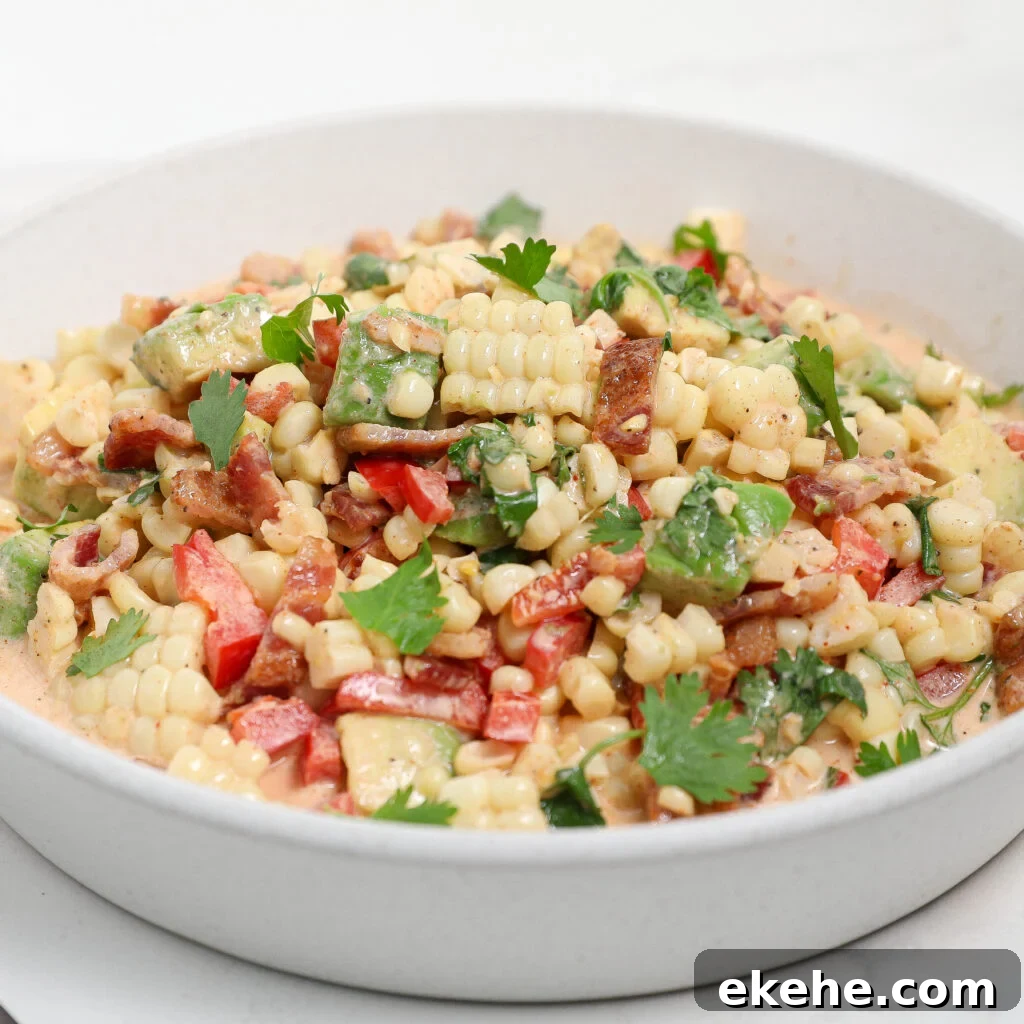 A white bowl filled with a vibrant corn and avocado salad with bacon, featuring corn kernels, diced red bell peppers, herbs, creamy dressing, and topped with fresh cilantro leaves.