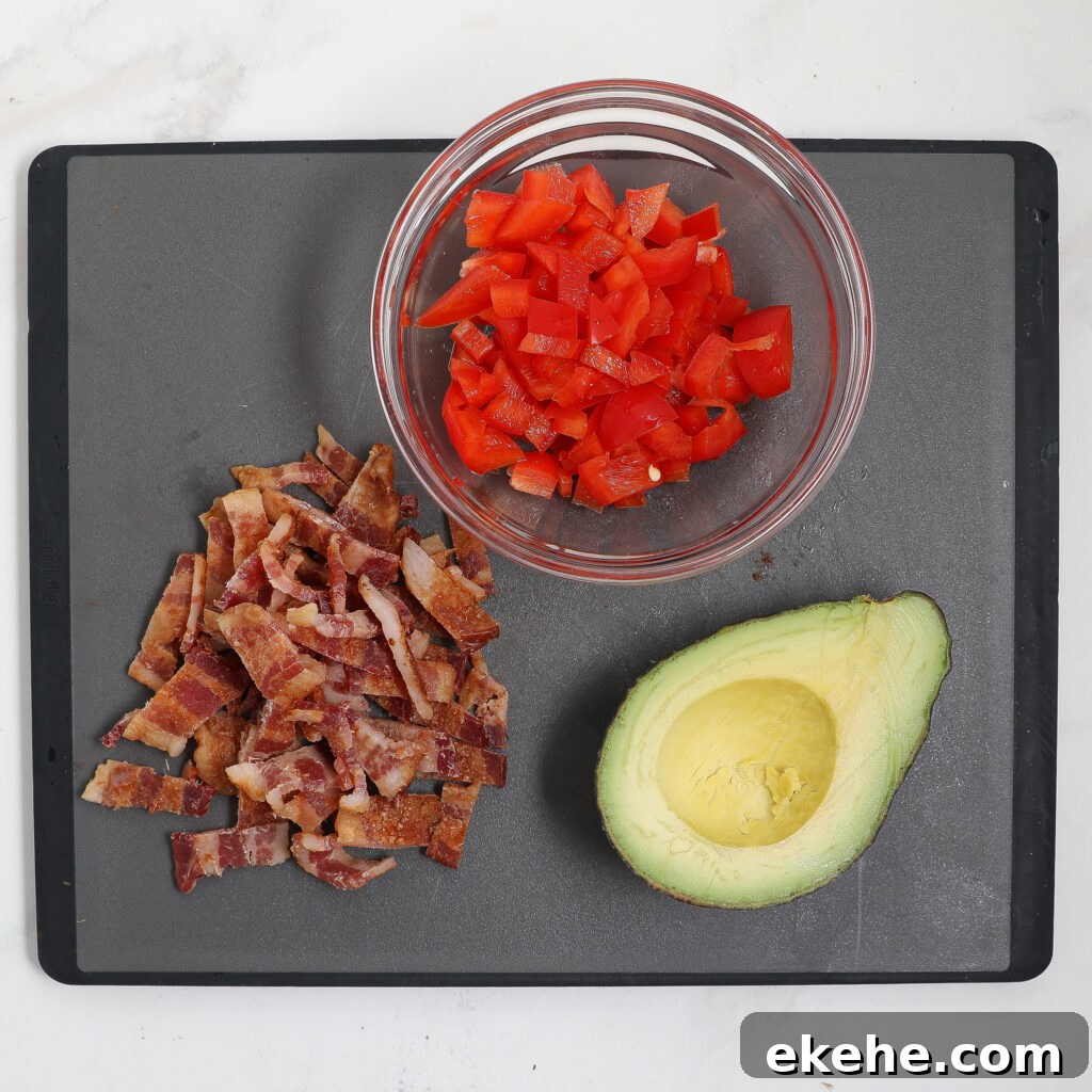 A black cutting board with chopped cooked bacon, a halved avocado, and a glass bowl of diced tomatoes on a white surface—perfect ingredients for a fresh corn and avocado salad with bacon.