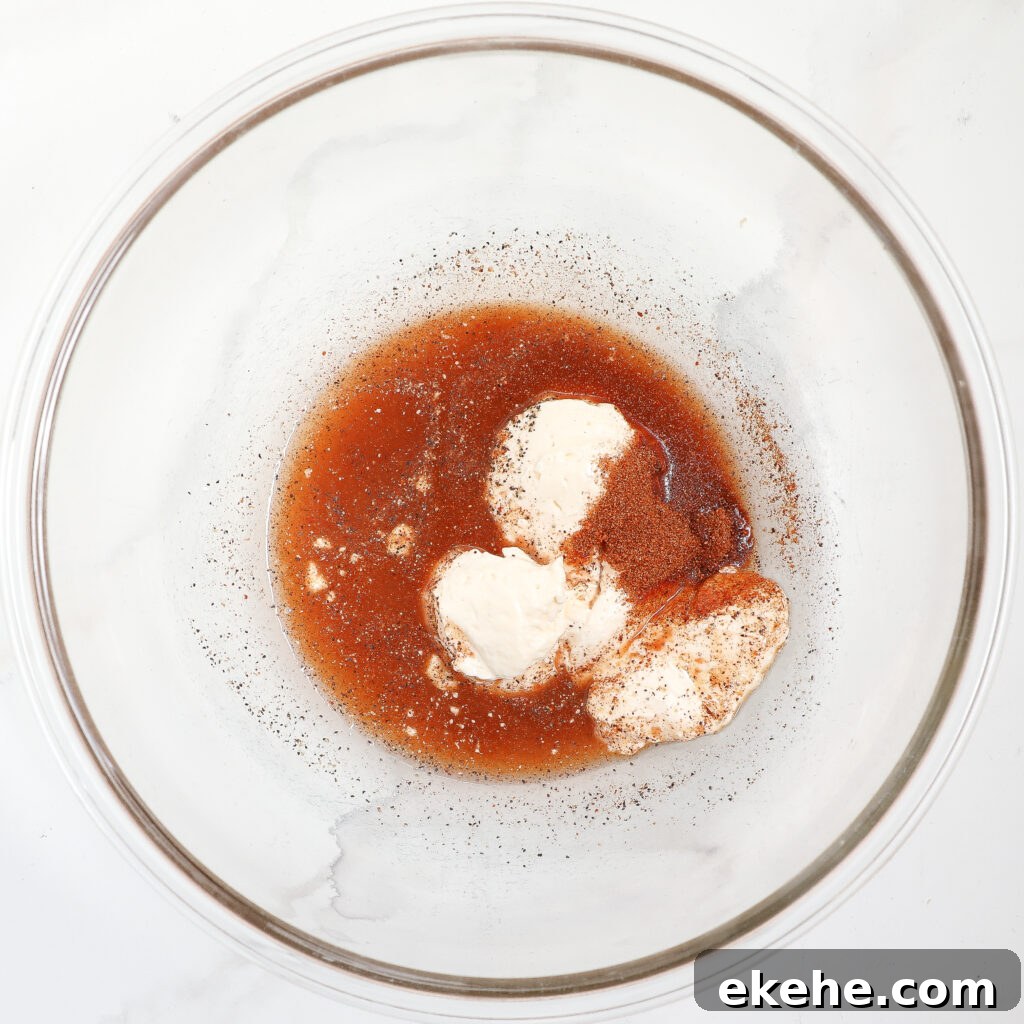 A glass bowl contains a mixture of mayonnaise, brown sauce, and seasonings—perfect for tossing with corn and avocado salad with bacon—set on a white marble surface. The ingredients are not yet fully blended together.