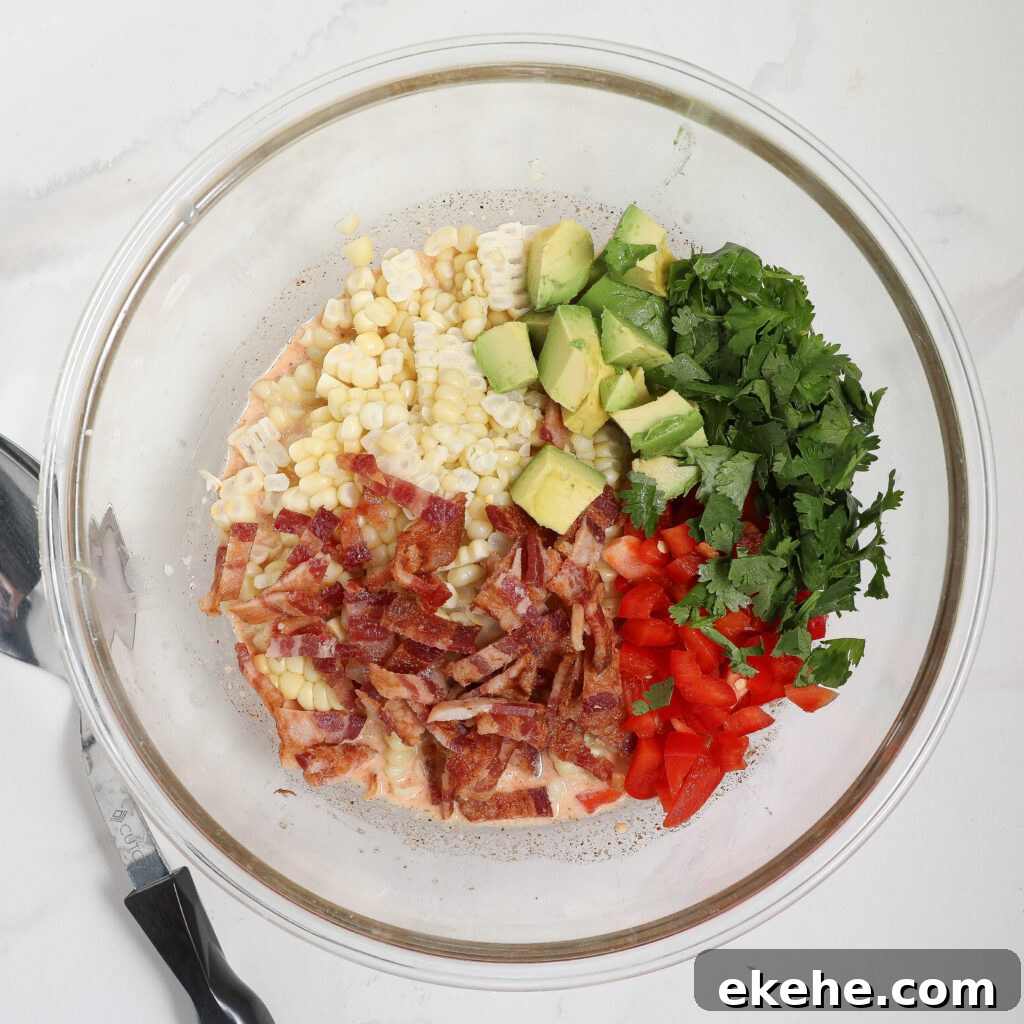 A glass bowl displays a corn and avocado salad with bacon, featuring chopped avocado, corn, cilantro, red bell pepper, and sliced cooked bacon arranged in separate sections on a white countertop. A spatula rests nearby.