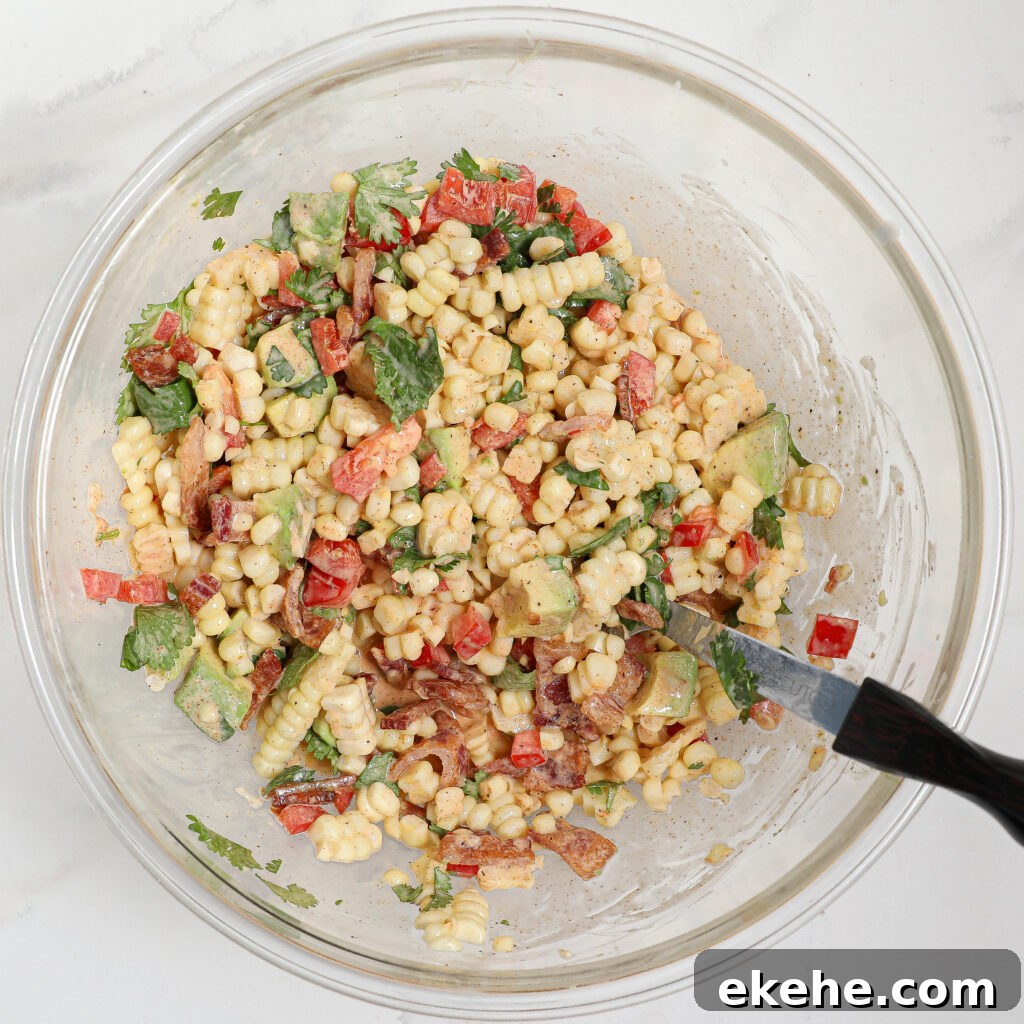 A glass bowl filled with a vibrant corn and avocado salad with bacon, featuring corn kernels, red bell peppers, cucumbers, chopped herbs, and a dressing, with a black-handled spoon resting inside.