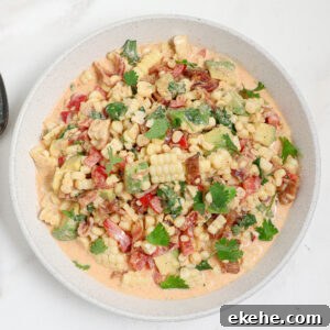 A bowl of creamy corn and avocado salad with bacon, chopped red bell pepper, cilantro, and a light orange dressing sits on a white surface.