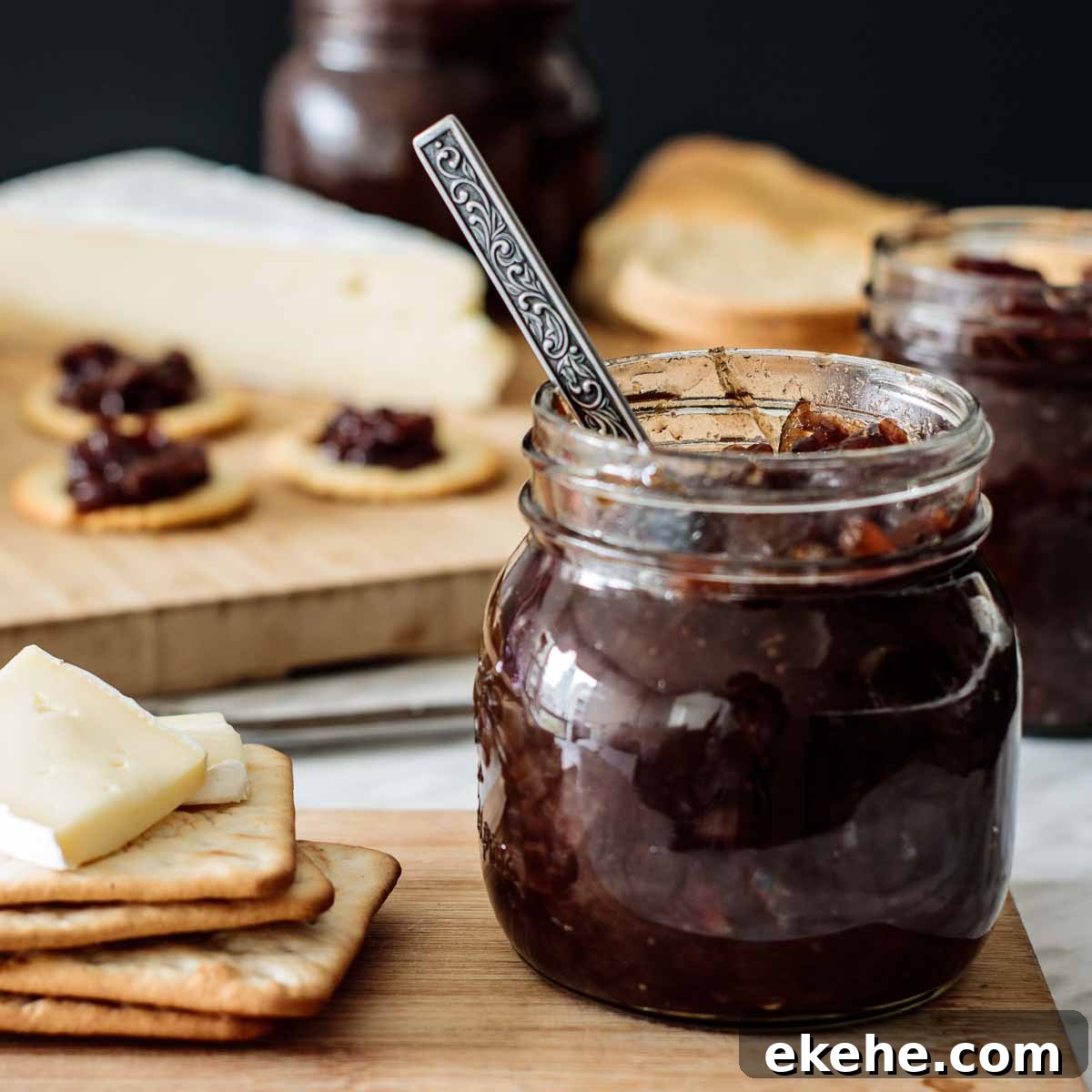A jar full of onion chutney with a spoon inside, and crackers with cheese on its side, over a wooden board.