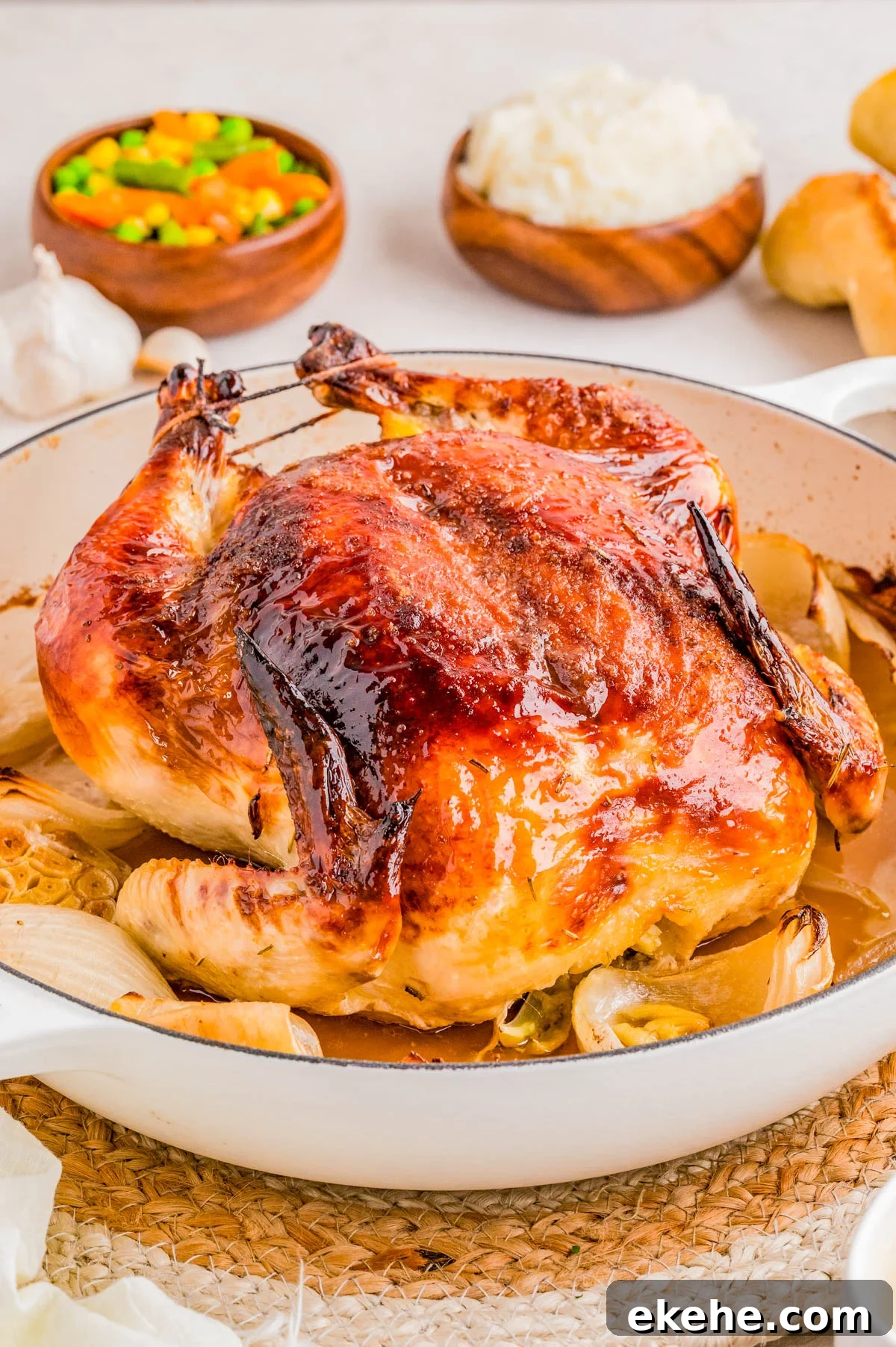Whole roasted chicken in a white dish with browned skin, surrounded by onions, with bowls of mixed vegetables, rice, and rolls in the background.
