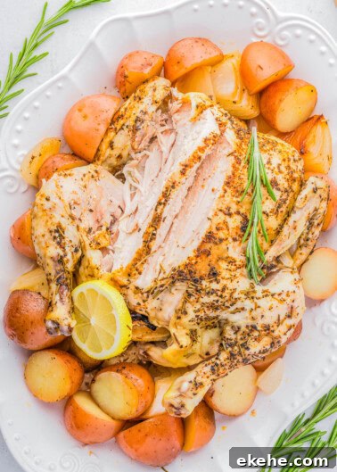 A roasted chicken on a white plate, partially sliced, surrounded by roasted potatoes and garnished with a lemon slice and sprigs of rosemary.