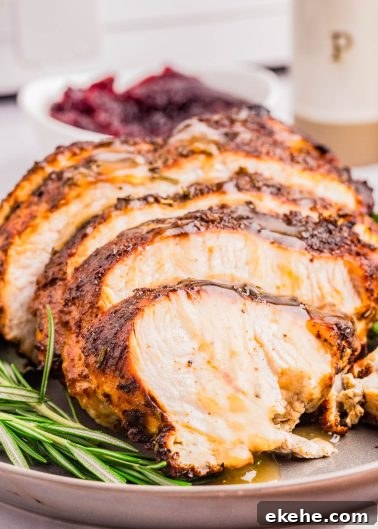 Sliced roasted turkey breast with herbs on a plate, garnished with rosemary, with a bowl of cranberry sauce in the background.
