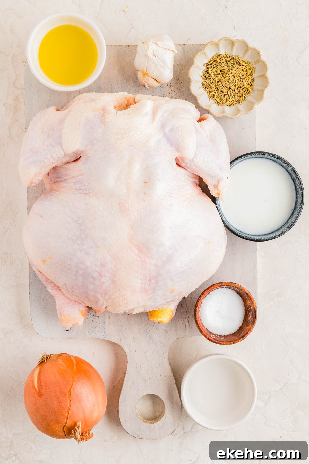 Raw whole chicken on a cutting board surrounded by olive oil, garlic, dried herbs, milk, salt, an onion, and a bowl of water.