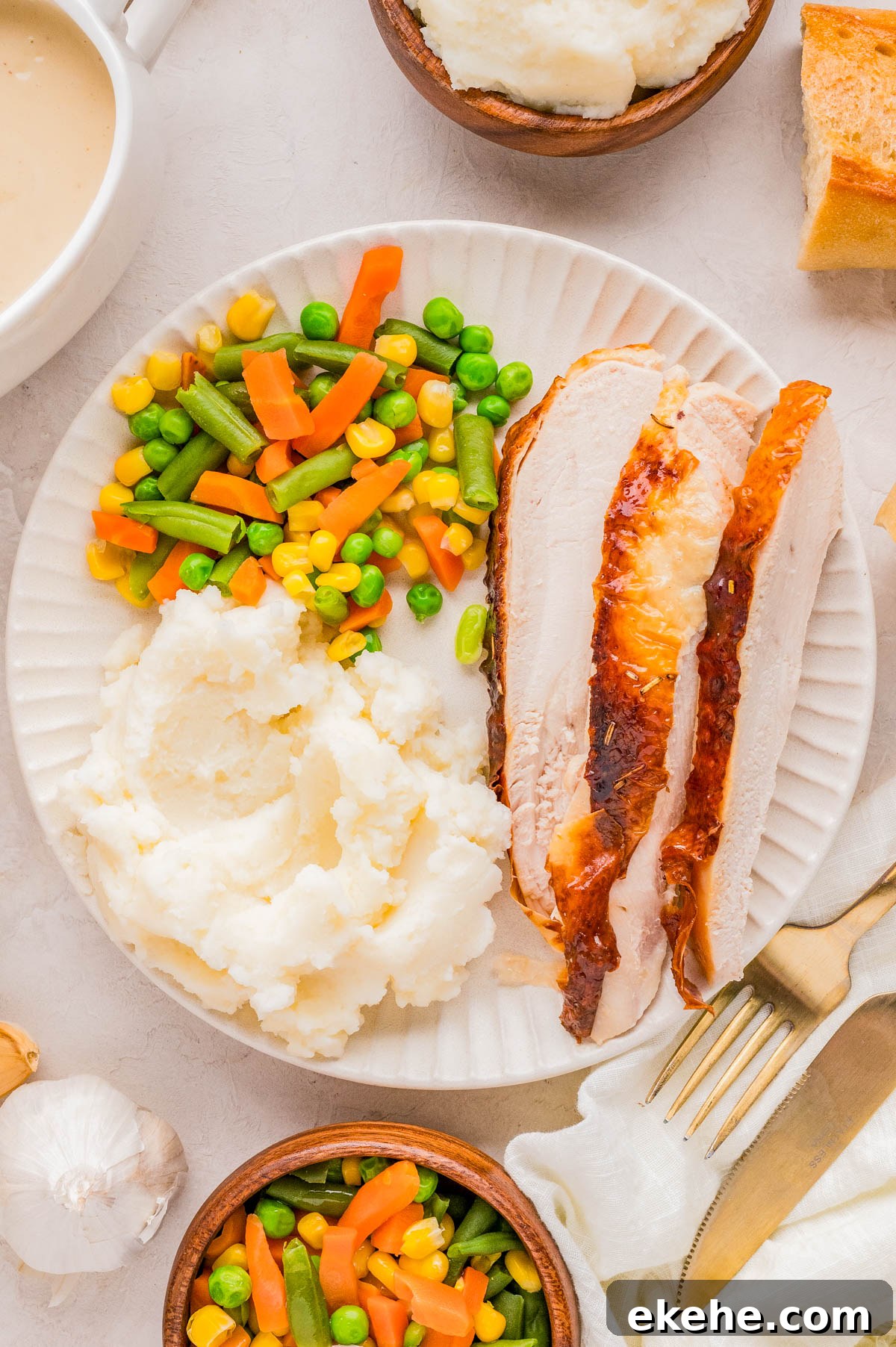 A plate with slices of roasted turkey, mashed potatoes, and mixed vegetables. Nearby are bowls of mashed potatoes and vegetables, a fork, knife, and bread.