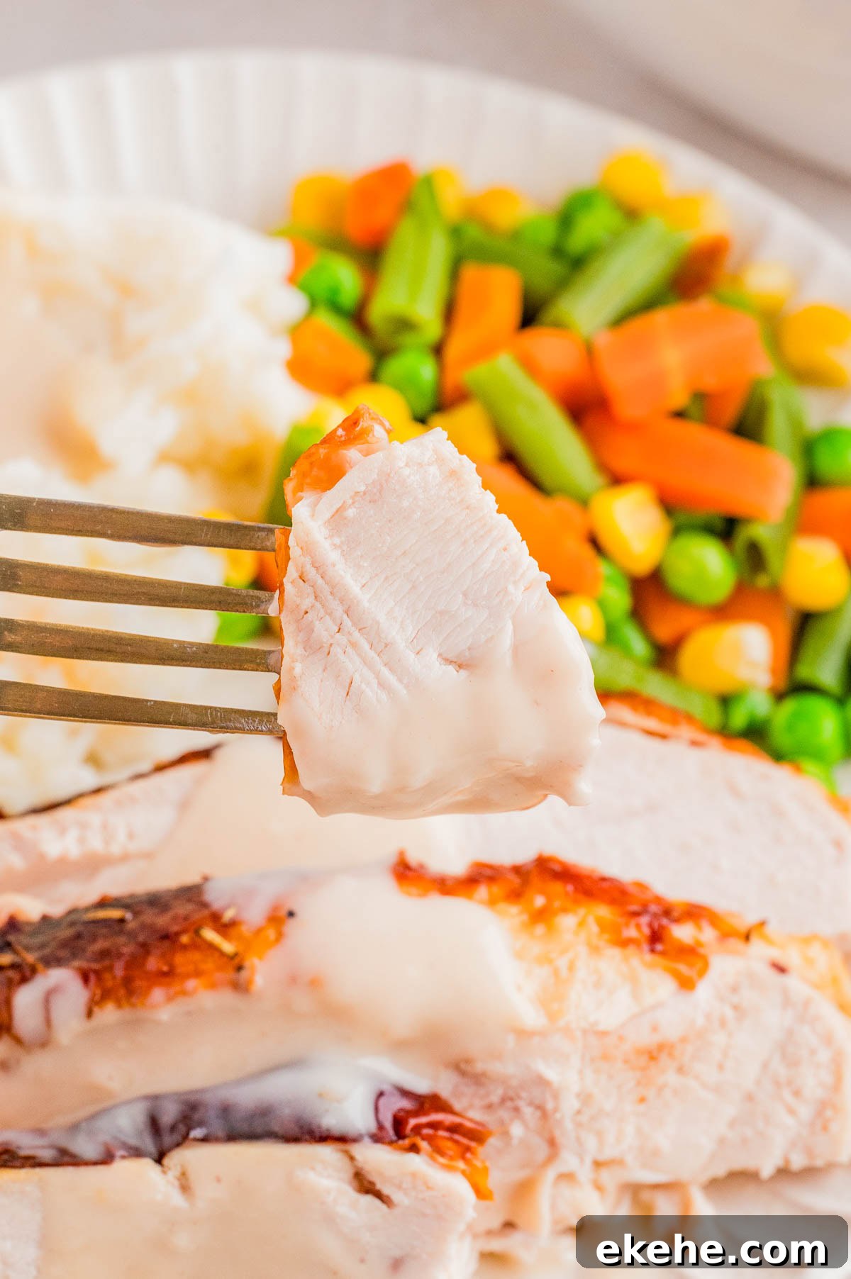 A fork holds a piece of turkey with gravy, with sliced turkey, mashed potatoes, and mixed vegetables on a plate in the background.