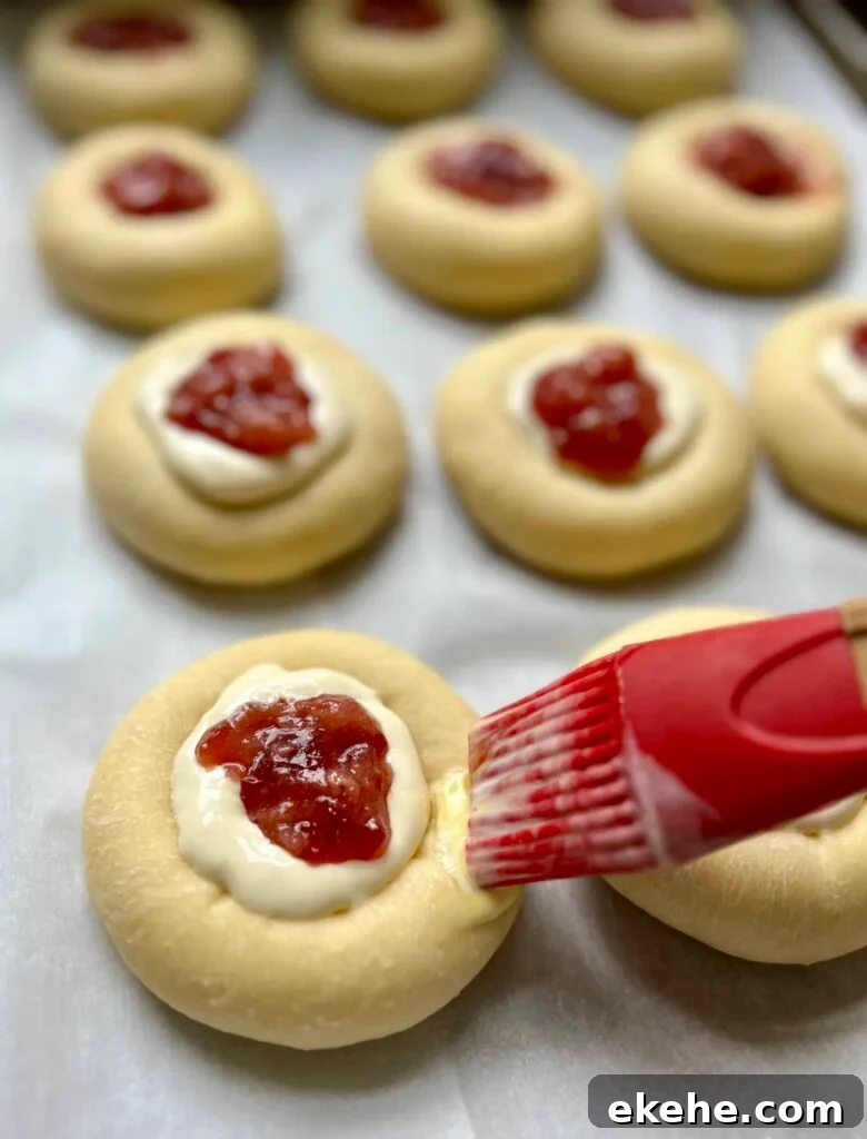 Brushing egg wash on sourdough kolaches filled with cream cheese filling and fruit jam.