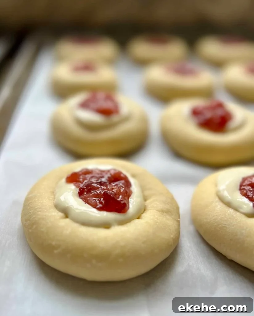 Unbaked sourdough kolaches with cream cheese and fruit jam filling on a baking sheet.