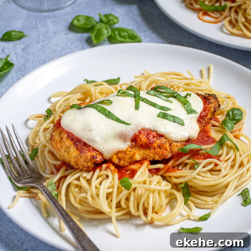 A white plate piled high with spaghetti topped with an Oven Baked Chicken Parmesan topped with melted mozzarella and garnished with pieces of fresh basil.