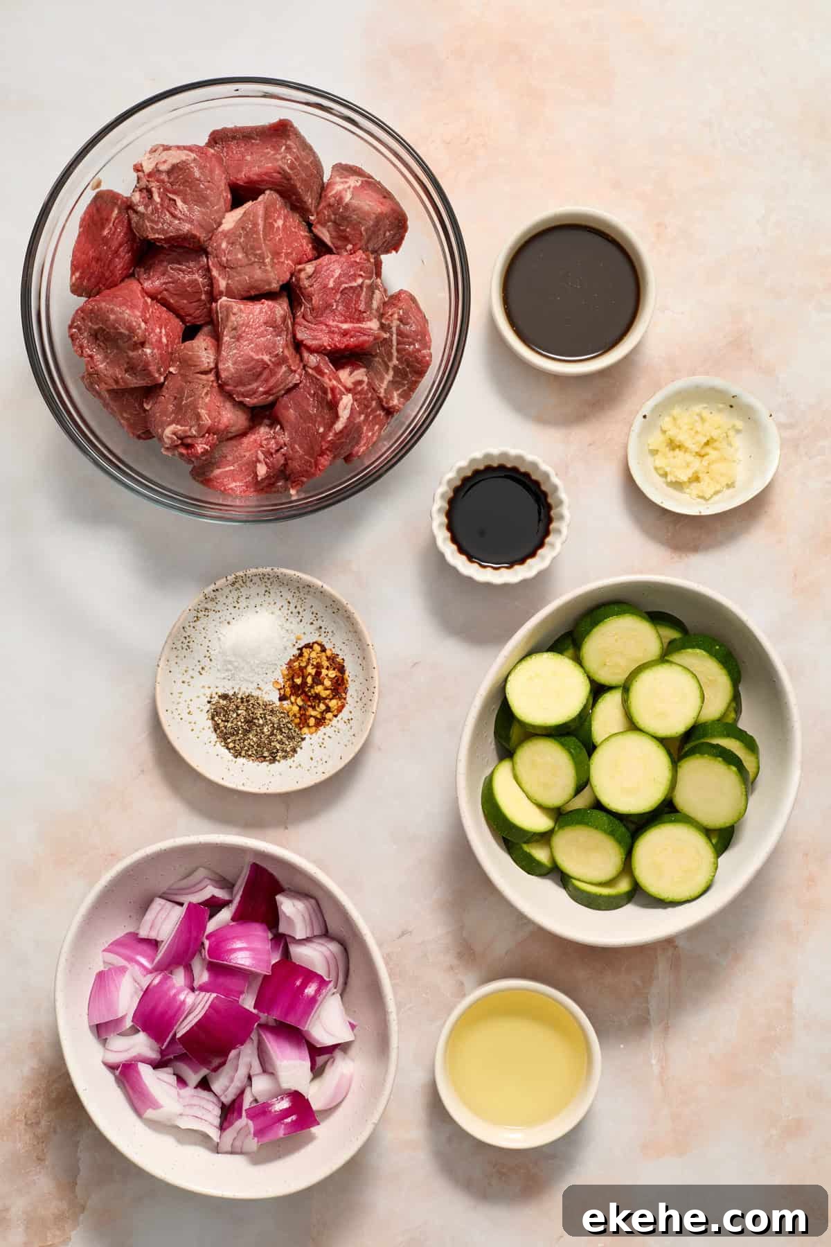 Sirloin steak cubed in bowl, sliced zucchini, sliced red onion and other ingredients arranged on surface.