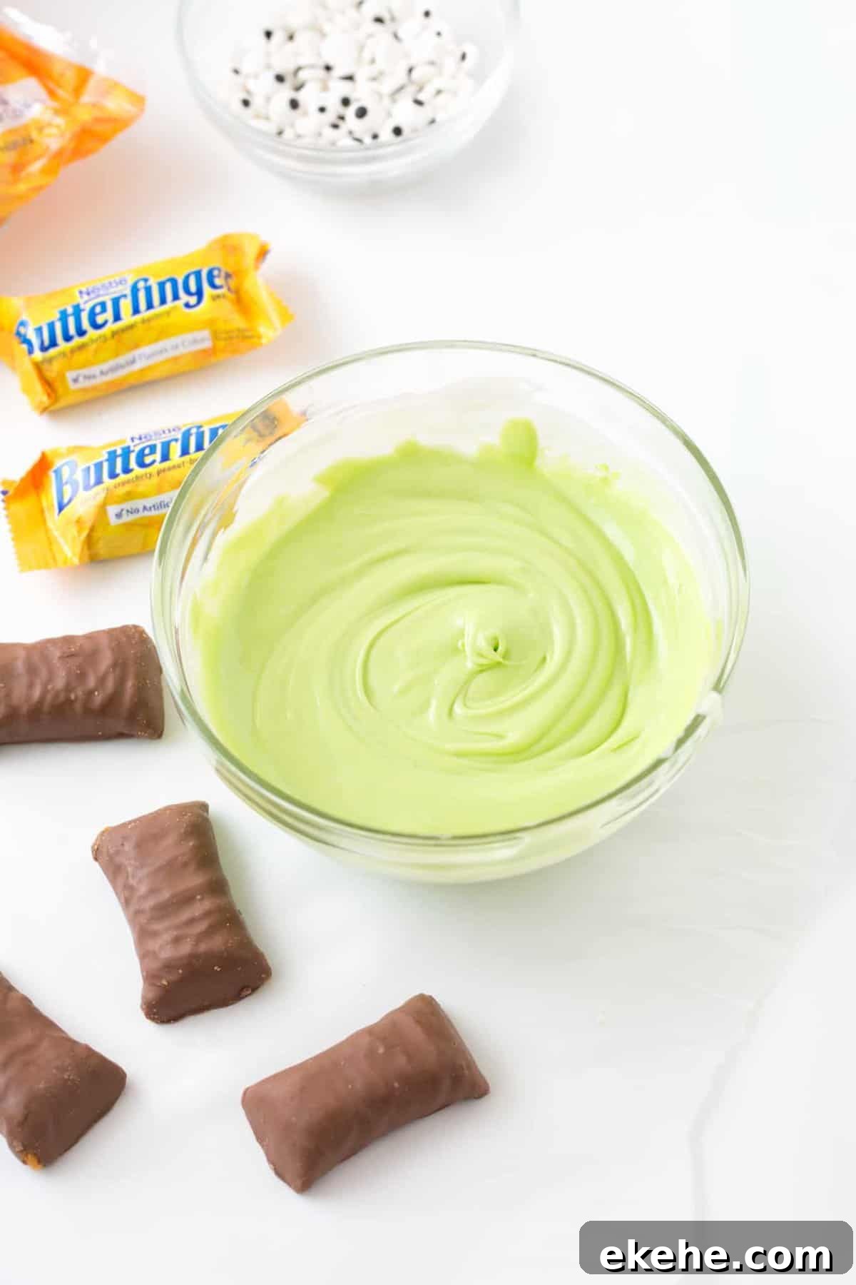 Bowl of melted green almond bark wth Butterfinger candy bars and a bowl of candy eyeballs off to the side.