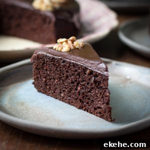 A slice of moist chocolate walnut cake with chocolate ganache and a walnut half served on a blue plate with the larger cake behind.