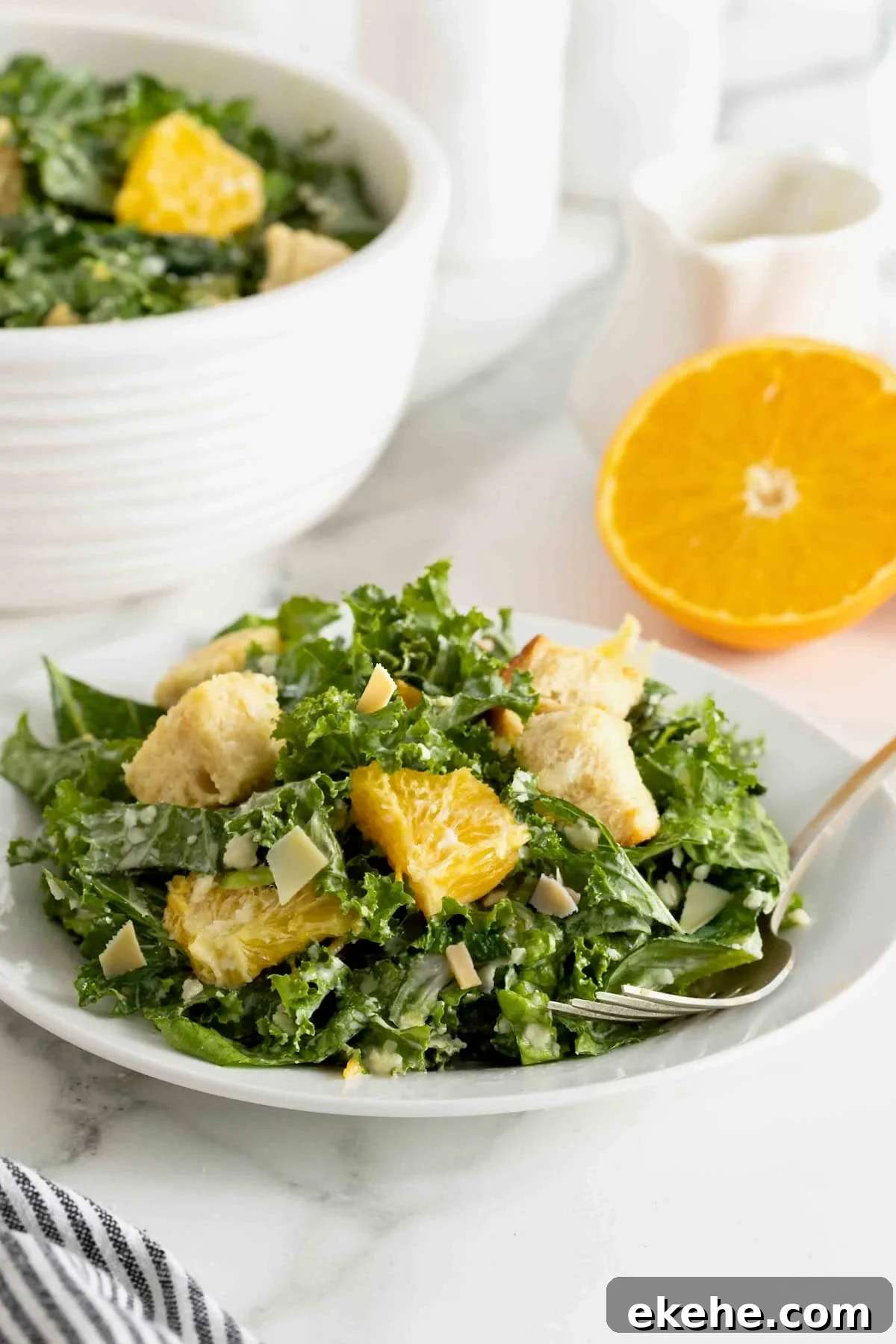 A white salad plate heaped with citrus caesar salad. There is a fork resting on the plate and a halved orange to the upper right.