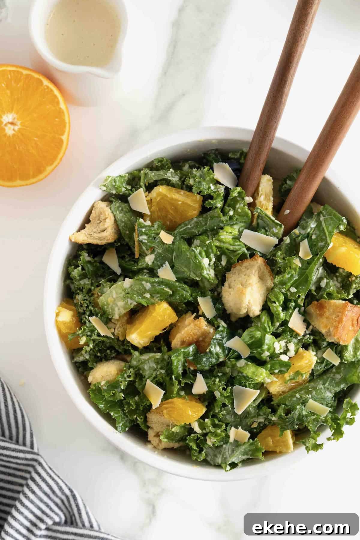 A white salad bowl of citrus caesar salad. Two wooden spoon handles are sticking out.