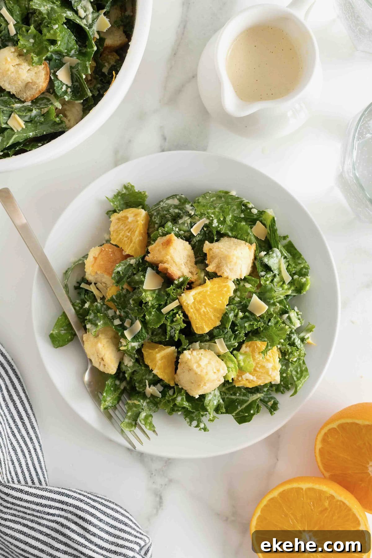 A white salad plate heaped with citrus caesar salad. There is a fork resting on the plate and a halved orange to the bottom right.