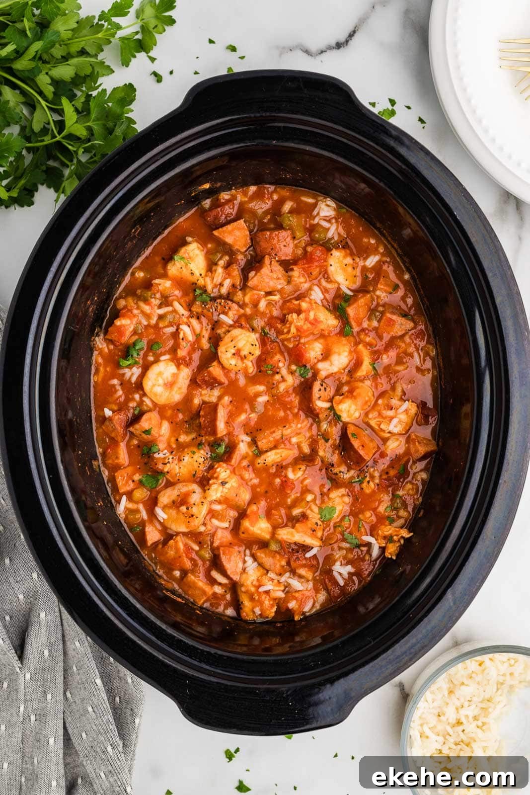 black slow cooker with slow cooked jambalaya surrounded by parsley, hand towel, white dinner plate and small bowl of cooked rice