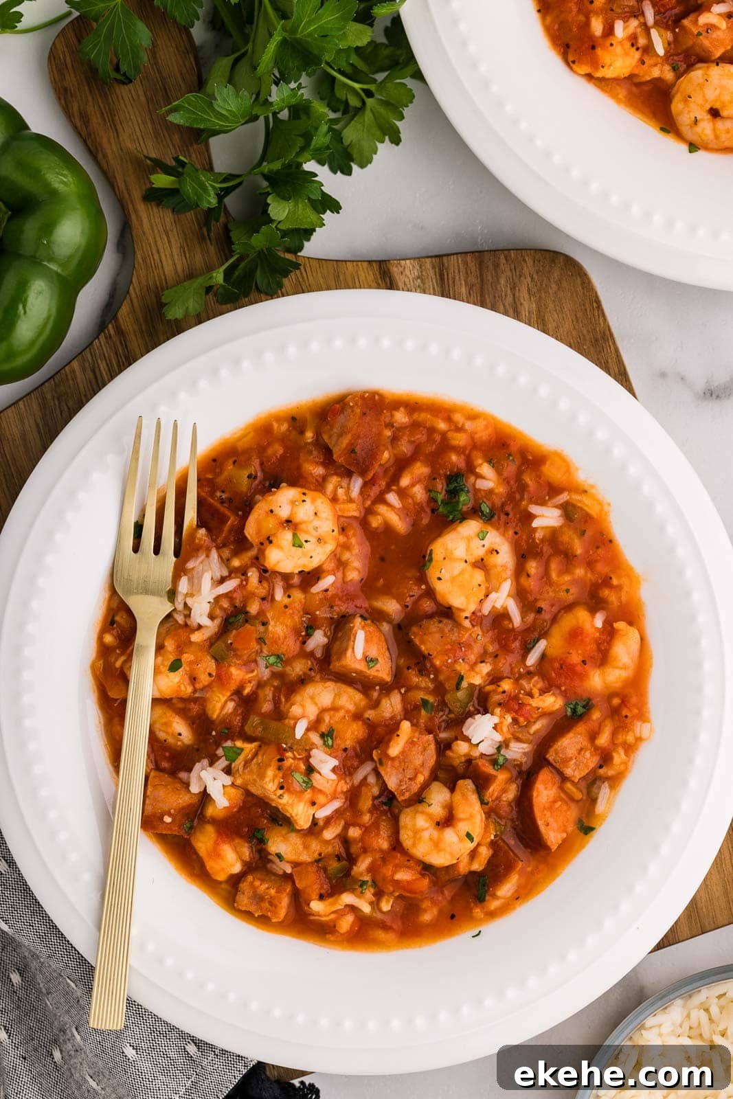 overhead view of white dinner bowl with jambalaya with fork surrounded by green pepper, parsley and another dinner plate