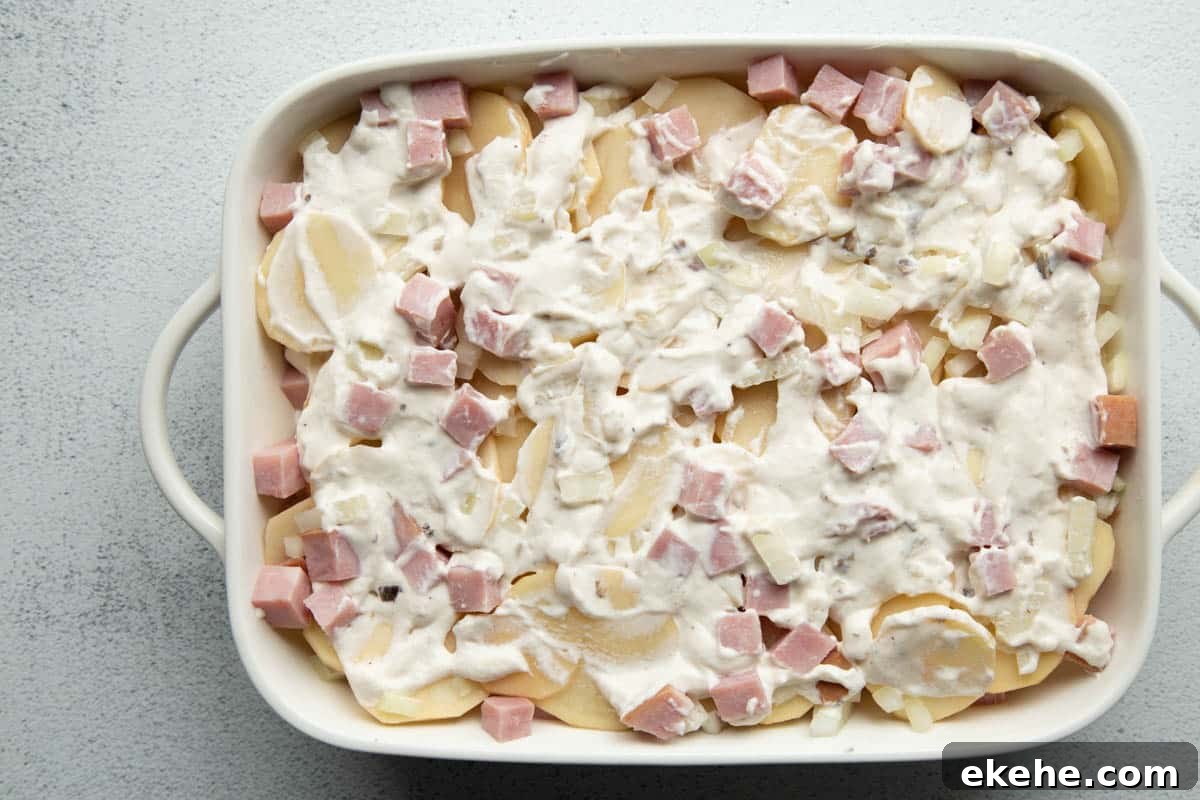 casserole dish with slices potatoes, diced ham, and a creamy sauce, shown unbaked.