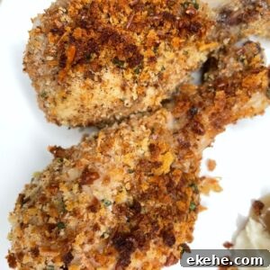 Two crispy, baked chicken drumsticks coated with a golden brown, crunchy breadcrumb and herb crust, served on a white plate. A small portion of mashed potatoes is visible at the edge.