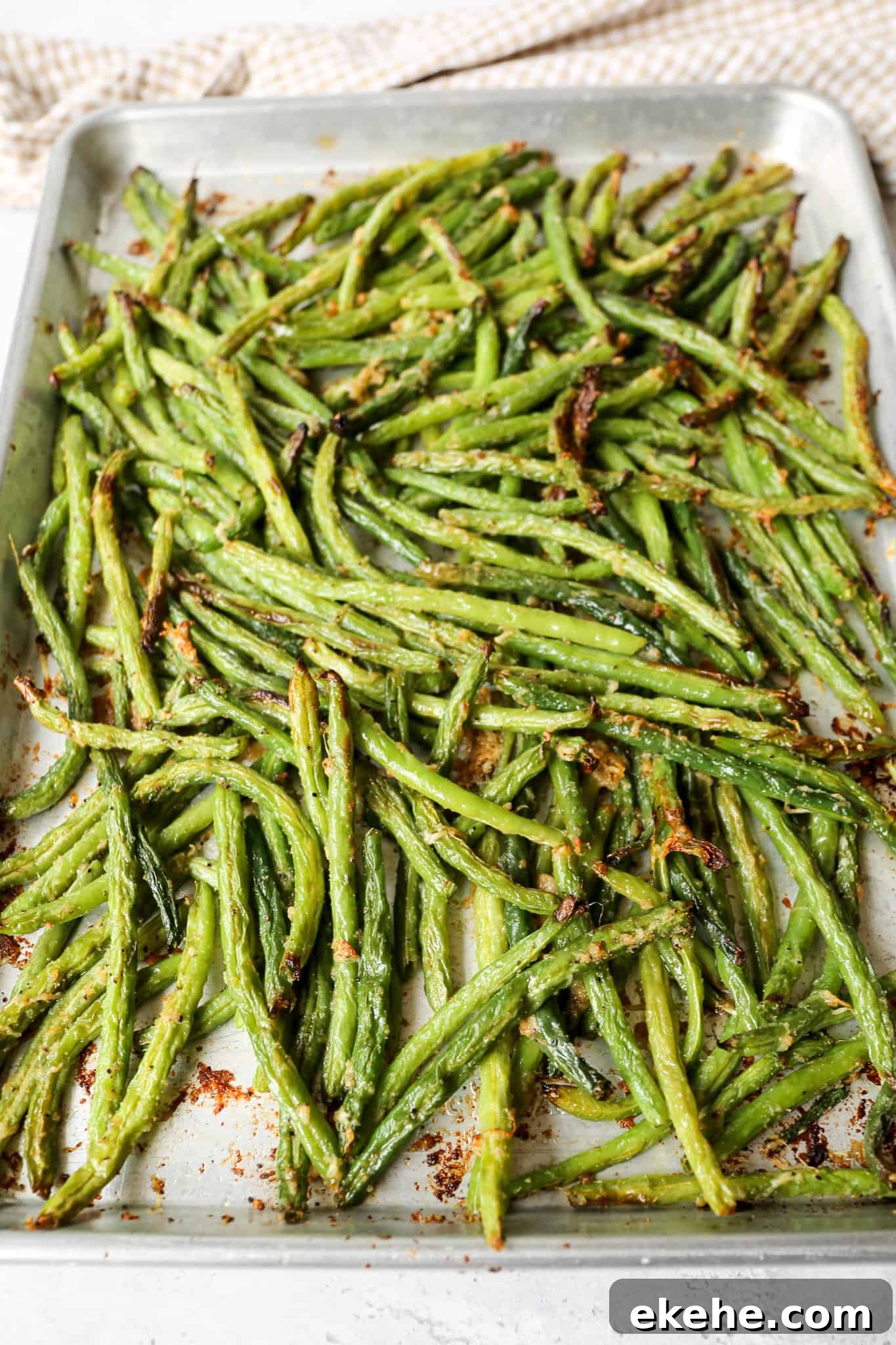a silver baking pan of oven roasted parmesan green beans next to a tan and white gingham kitchen towel