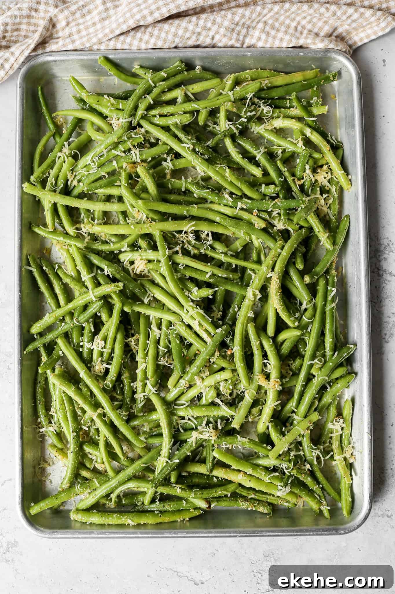 a silver baking pan of oven roasted parmesan green beans next to a tan and white gingham kitchen towel
