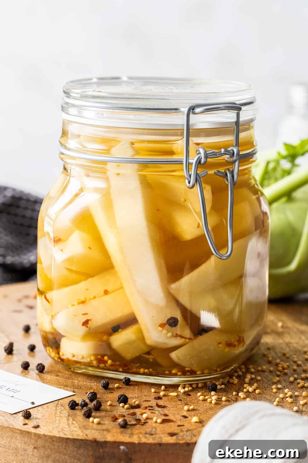 Jar of Pickled Kohlrabi, sitting on a round wooden board, with some herbs and spices around the edge.