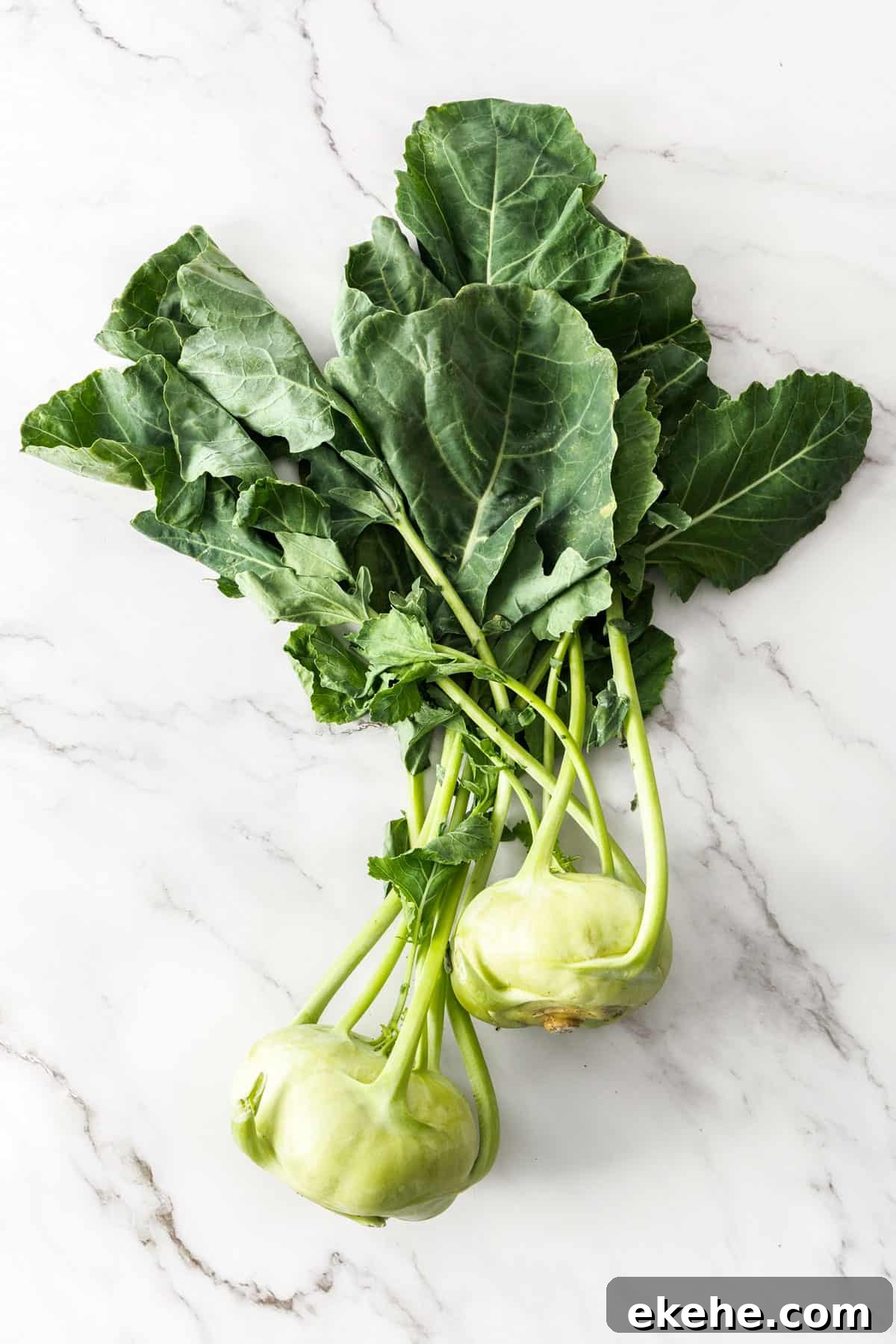 Two whole kohlrabi, sitting on a white marble background.
