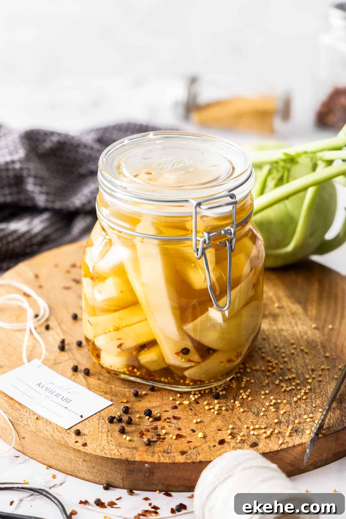 Jar of Pickled Kohlrabi, sitting on a round wooden board, with some herbs and spices around the edge.