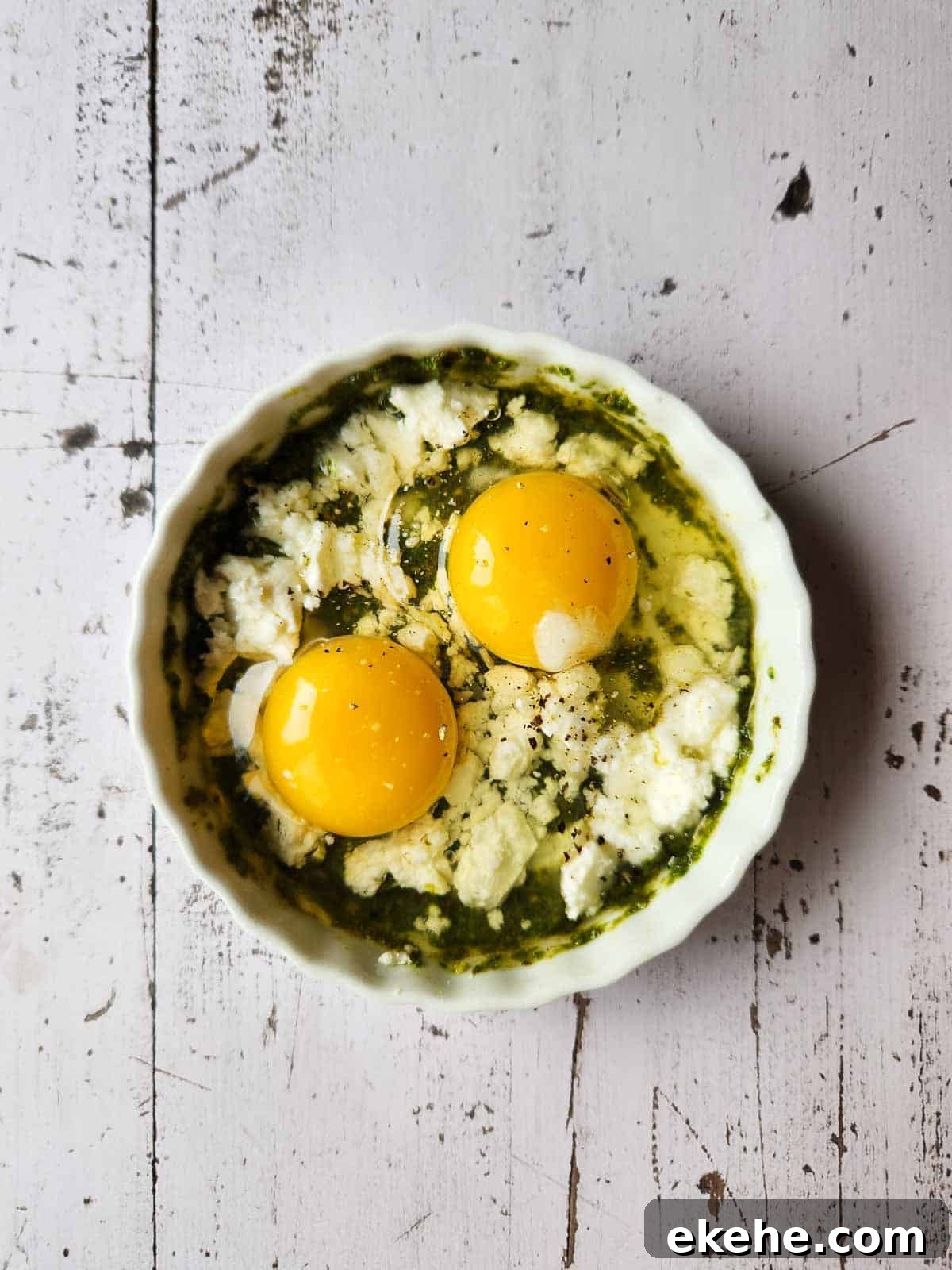 Pesto, feta, cream, and two eggs in a white ramekin.