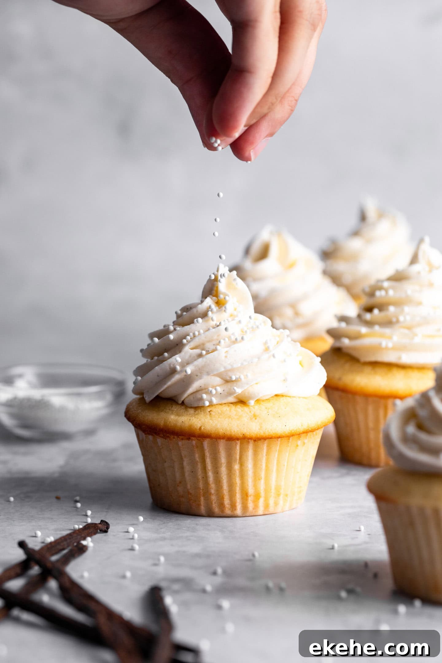 Sprinkles being dropped on top of vanilla cupcakes with vanilla frosting swirled on top.